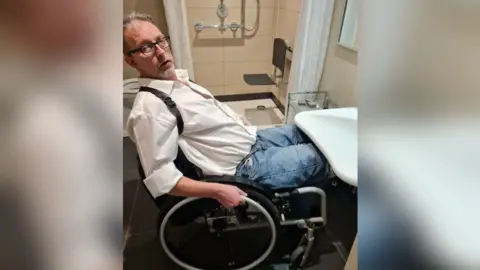 Rohan Tarry Rohan Tarry is in his wheelchair trying to access a sink but he cannot get close enough to it to reach it. He is looking at the camera. Behind him is a bath with a disabled seating platform over it