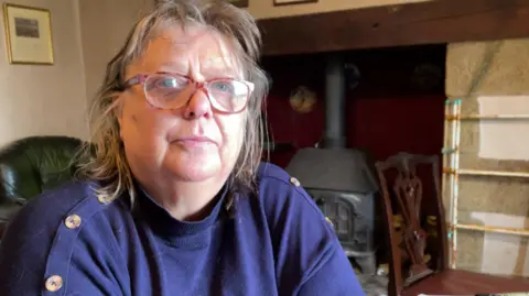 Marie Wills is sitting on a chair in front of a log burner. She is wearing a navy jumper and is looking at the camera.
