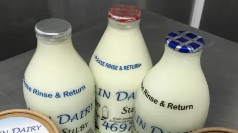 Aalin Dairy Three milk bottles full with white milk, with foil tops. One is silver, another is red, the third is blue.
