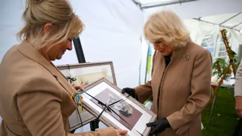 PA Media Camilla wears a brown jacket with a glittery broach and holds a portrait of a small dog. Another woman in a brown jacket also holds the portrait.
