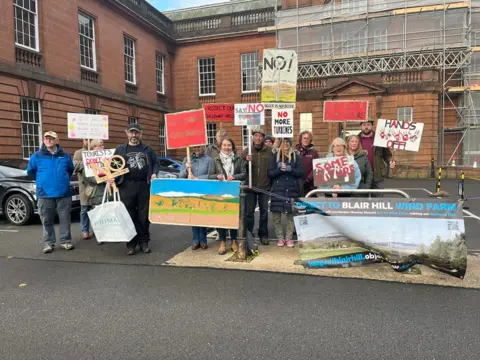 A group of wind farm protesters with placards stands outside Dumfries and Galloway Council HQ