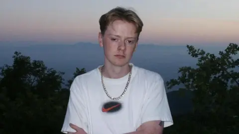 Olivia Osby A man with red parted hair stands outside and poses for a picture during sunset. He is wearing a white top, a gold chain, and a gold watch. The top of a tree line can be seen in the background, as can hills faintly in the distance. The sky is a muted blue-grey colour with some dull orange. 