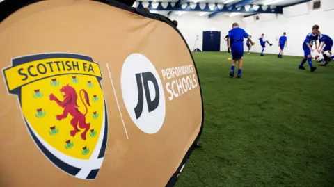 SNS A banner advertising performance schools with the SFA logo. Boys in blue football strips are playing football alongside it in an indoor area.