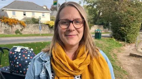 BBC Beth is looking at the camera while standing on grass. Behind her is a pram, a road and a large house. She is wearing a light blue denim jacket and a mustard-coloured scarf.