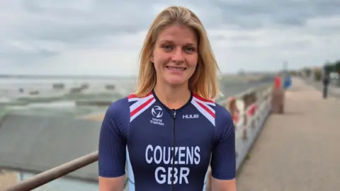 Stuart Woodward/BBC Georgia Couzens has shoulder-length straight blonde hair. She is wearing a navy blue skin-tight top with her surname and the letters GBR written in bold white font on the front, as well as a logo for the World Triathlon Championships. There are red and white detailing to her shoulders. Georgia is stood on Southend seafront with beach huts in the background.