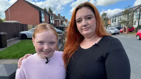BBC Toni and her ten-year-old daughter Olivia. Toni, wearing a black top with ginger hair, has her arm around her. Olivia is wearing a pink jumper and is smiling. They are standing on a residential street. 