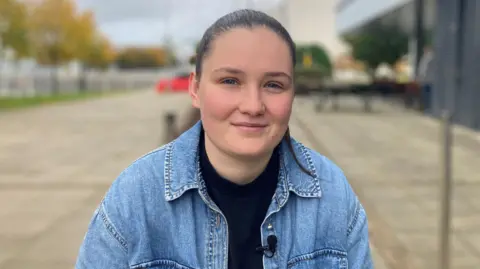 BBC Andrea Bisset, a fresh-faced young woman, wears a denim jacket and has her hair tied back. She smiles at the camera, a large outdoor paved space behind her.
