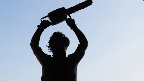 Getty Images A silhouetted image of somebody dressed as Leatherface holding a chainsaw above their head