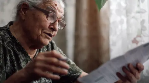 Getty Images Older woman wearing glasses looks at a bill with curtains and net curtains behind her.