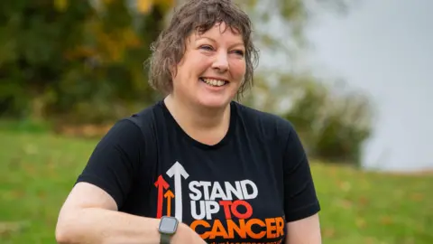 Supplied Caroline Palmer wearing a black Stand Up To Cancer t shirt and leaning on a wooden post.