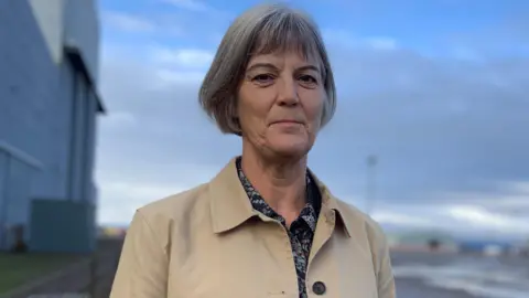 BBC Julia Longbottom, a lady with short grey hair stares at the camera with a serious  expression. She is wearing a patterned blouse and a beige rain jacket. An industrial complex can be seen in the background.