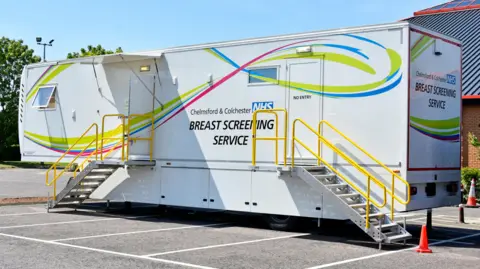 Alamy Mobile breast screening van situated in a car park. It is rectangular. Steps lead to the entry and exit doors. The livery is white with coloured patterns alongside the logo of an NHS trust. 