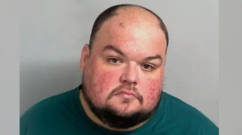 Essex Police Gavin Plumb looks serious, if not slightly angry, in a picture taken in police custody. He has a chinstrap beard and a moustache. He is wearing a green T-shirt.
