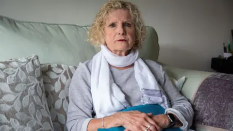 BBC Sian Jones at home - a woman with a curly blonde bob, picture sitting of a grey sofa, holding a hot water bottle across her tummy.
