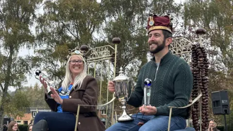 Aimee Dexter/BBC A woman and a man sitting on chairs that say Queen Conker and King Conker across the back. They are both holding silver trophies, wearing crowns and smiling. She has long grey hair and is wearing glasses, a brown coat and blue jeans. He has a dark beard and is wearing a green jumper and blue jeans.