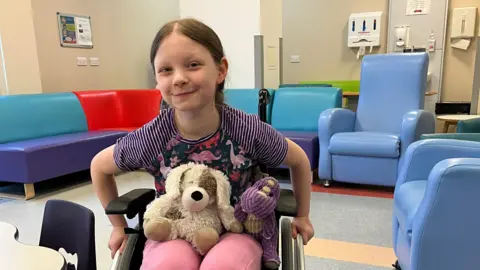 Cancer Support UK Ivy is sitting in a wheelchair in what looks like a hospital waiting area and smiling at the camera. Two teddy bears sit on her lap. Behind her is a multi-coloured L-shaped vinyl sofa and a couple of blue very high-back armchairs.