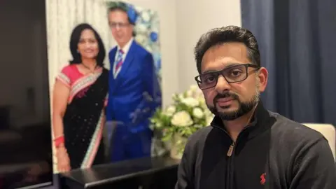 Chrissie Reidy/BBC A man, Miten Patel, in a dark jumper, looking into the camera. In the background is a large image of two people in formal attire and a bouquet of white flowers. 