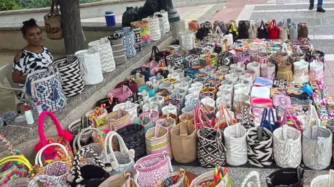 BBC A woman selling tens of mochilas on the street in the Colombian city of Riohacha, with the colourful bags laid out in front of her