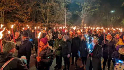 BBC Participants of the torchlight parade walk through the streets of Lincoln. The majority of people are wearing warm, winter clothing and are carrying long candles which are burning. Trees are visible in the background.