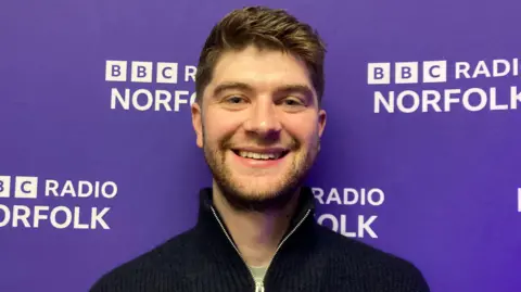 Alex Arnold/BBC Alex Green, a 26-year-old man who is standing inside the BBC Radio Norfolk studio. He is standing in front of a purple wall which has BBC Radio Norfolk branding on it.