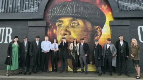 Derek Brennan A group of people dressed in flat caps and tweed suits. stand in front of a large colourful mural of Thomas Shelby.