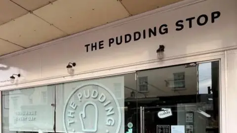 Simon Dedman/BBC The Pudding Stop, in St Albans, closed, with benches outside and a car to the left. The door is shut, with blinds half way down on the window. 