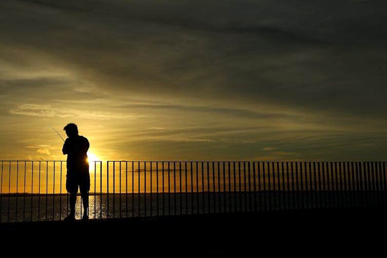 A man stands against a barricade fishing, with the sunrise behind him.