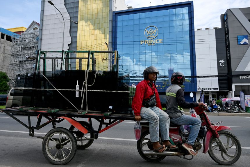 Workers ride their motor-cart loaded with glass past a branch of the Prince Bank in Phnom Penh on October 15, 2025.