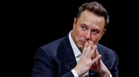 Reuters Elon Musk wearing a blue suit and white shirt with his fingertips touching and resting on his lips against a black background