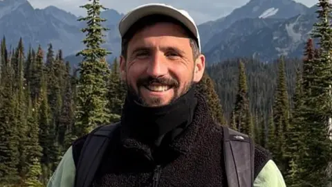 Oliver Gooch Oliver Gooch standing smiling in front of trees and mountains smiling. He has dark hair and a beard and is wearing a white baseball cap and black fleece and scarf
