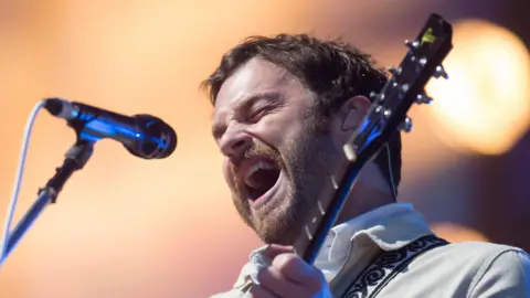 PA Media Kings of Leon lead vocalist Caleb Followill closes his eyes and sings into a microphone. He is wearing a light-coloured shirt and also playing a guitar.