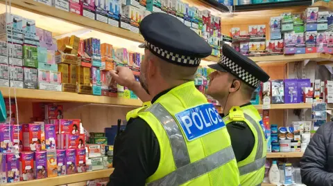 National Crime Agency Two male police officers, wearing police caps and high-vis police branded vests, examine vapes on a shelf at a store. 