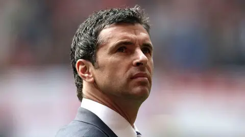 Getty Images Gary Speed looks upwards during a 2011 match between Wales and England in the stadium. He is wearing a dark suit and tie.