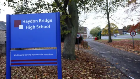 Iain Buist/NCJ Media A blue sign that reads in white lettering, 'Haydon Bridge High School' on a patch of grass covered in brown leaves. It stands in front of a tree. To the right is a path leading to a school. 
