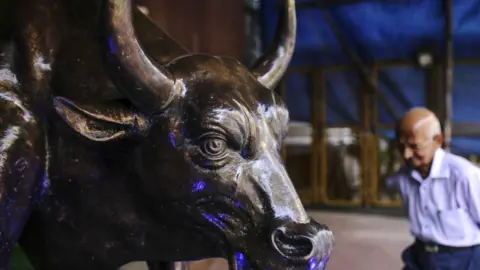 Bloomberg via Getty Images A bronze bull statue stands at the entrance to the Bombay Stock Exchange, with a balding man walking past it. 
