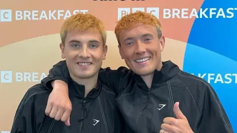 jord_adams Cian Adams and brother Jordan smile. They are wearing black tracksuit tops and have dyed orange hair. They are standing in front of a BBC Breakfast backdrop sign.