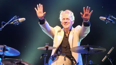Getty Images A white-haired man wearing a white suit and black shirt holds his hands in the air while sat at his drumkit. He is illuminated by stage lights and is smiling for the crowd.