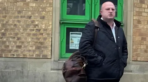 Brian Farmer/BBC Michael Kempson: A bald man standing in front of brown-brick wall. He has his hands in the pockets of a black, zipped, jacket and is carrying a brown bag over his right shoulder.

