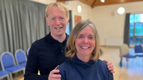 BBC/Seb Cheer A man and woman in their 50s stand and smile at the camera, in a village hall.