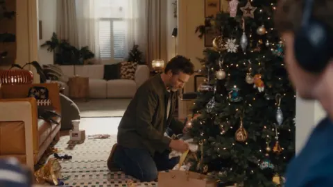 John Lewis A father wearing a khaki shirt, a white t-shirt and blue jeans opening a present under a Christmas tree decorated with classic baubles. His son is watching, wearing a blue jumper and black headphones. 
