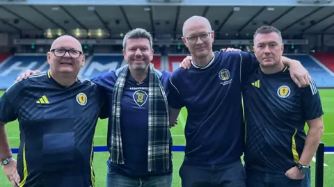 BBC Four male Scotland fans, all in navy blue Scotland jerseys, stand arm-in arm with the pitch and the seating stand behind them at Hampden Park