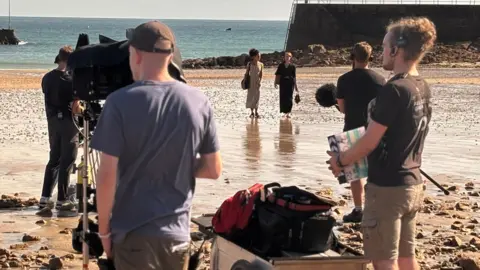 BBC A film crew shoot a scene for the second series of Bergerac on the beach at St Brelade's Bay in Jersey. The sea is in the distance. Two actresses are walking along the sand.