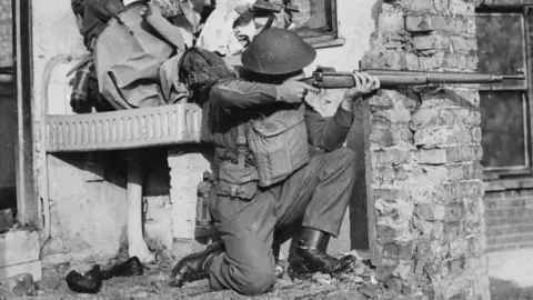Getty/Fred Rammage A black and white image of a soldier crouching down beside a wrecked building. He points a Lee Enfield rifle, his eye lining up the sights, finger on trigger.