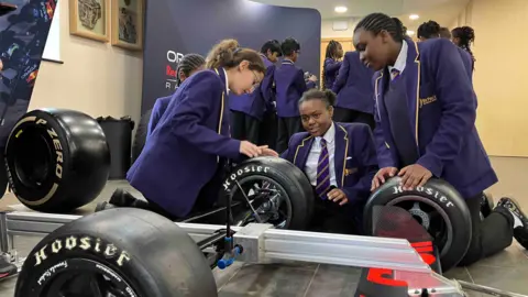 Sam Read/BBC Children in purple school uniform carrying out a mock pit stop on a frame with racing car wheels attached.