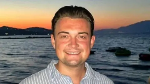 Family Photo A white young man with brown hair, wearing a striped blue and white shirt. He's stood in front of a body of water with a sunset scene in the background.
