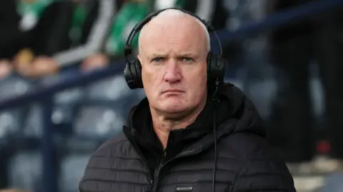 SNS Kenny Macintyre in a football stadium wearing a black puffer jacket and black headphones