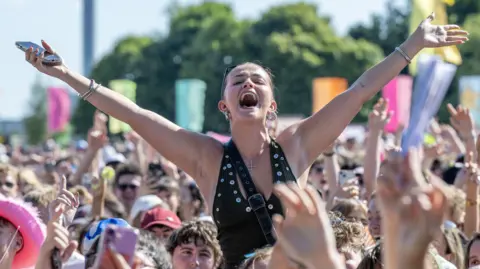 PA Media A crowd shot of TRNSMT. One woman is on someone's shoulders and has her hands in the air. People in the crowd have their phones up or their arms in the air. 