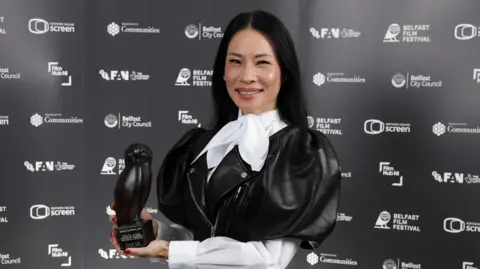 PRESS EYE Lucy Liu smiles while posing with her award at the Belfast Film Festival.  She has long, straight, black hair and is wearing a black leather frock coat with puffed sleeves over a white blouse tied with a large bow at her neck.  She is holding an award shaped like an owl. 
