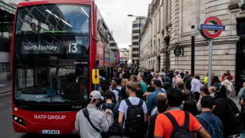 Getty Images Commuters queue for buses outside Victoria underground train station. It is a busy day with a sea of people outside the bus. A red North Finchley 13 bus has arrived at the stop. 