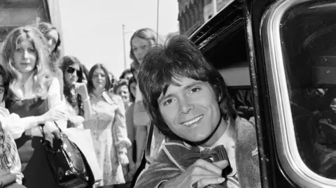 Getty Images A black and white image of Sir Cliff Richard leaning out of a Mini. He is wearing a dinner suit and bow tie. There is a crowd of women behind the vehicle looking towards the camera.